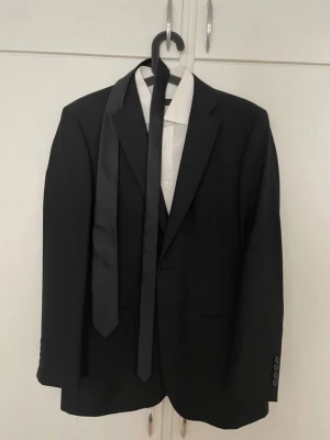 Svart kostym från Dressmann  - Klassisk svart kostym sett från Dressmann i storlek L, med matchande svart slips och vit skjorta samt byxor.  Kavajen har enkelknäppning, smala slag och tre knappar vid ärmslutet. Använd 1 gång