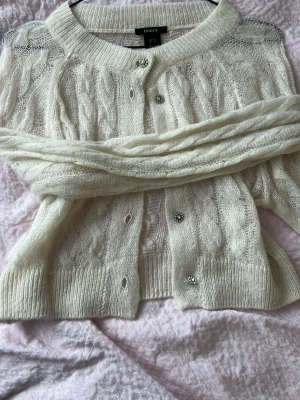 Vit kabelstickad kofta från Lindex  - Mysig vit kabelstickad kofta från lindex med rund halsringning och dekorativa knappar. Koftan har långa ärmar och ribbade muddar. Tillverkad i mjukt, fluffigt material som ger en lyxig känsla.