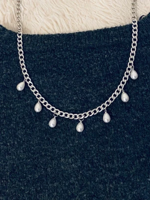 Silverfärgat halsband med pärldroppar - Snyggt halsband i silverfärgad rostfritt stål med sju droppformade vita pärlor som hänger längs kedjan. Kedjan har en chunky design som ger ett trendigt intryck och pärlorna ger en elegant touch. Perfekt accessoar för att lyfta din outfit.