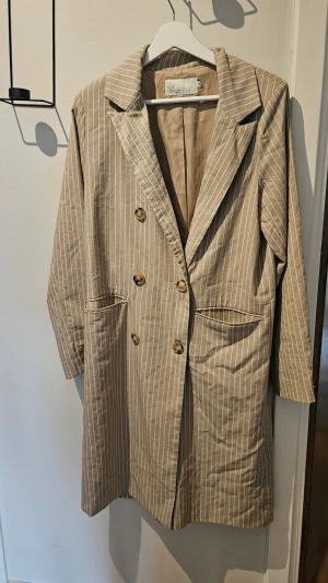 Beige randig kavaj från Nelly Trend - Snygg beige kavaj med vita vertikala ränder från Nelly Trend. Dubbelknäpp framtill och två fickor. Lätt oversized passform och klassisk krage. Perfekt för en stilren look.