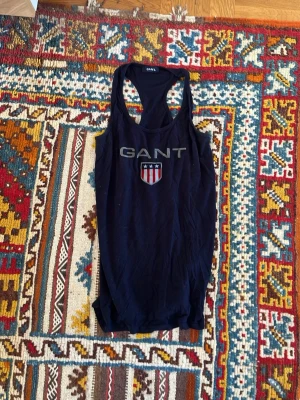 Mörkblå linnetopp från GANT - Snygg mörkblå linnetopp från GANT med klassiskt tryck och logga framtill. Modellen är ärmlös med brottarrygg och har en sportig vibe. Perfekt till sommaren och enkel att matcha med jeans eller shorts.