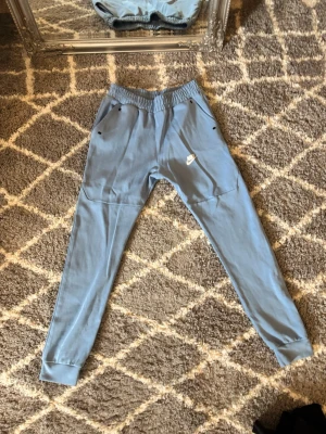 Blå Nike mjukisbyxor med fickor - Snygga ljusblå mjukisbyxor från Nike med resår i midjan och muddar vid bensluten. Byxorna har två sidofickor med dragkedja och en vit Nike-logga på låret. Perfekta för chill eller träning.