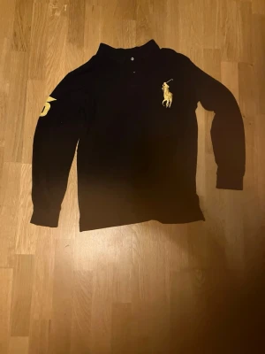 Svart långärmad tröja från Ralph Lauren - Svart långärmad tröja från Ralph Lauren med klassisk gul broderad logga på bröstet och siffran 3 i gult på ena ärmen. Tröjan har krage och knäppning vid halsen, perfekt för en stilren och sportig look.Den är custom fit men är typ nånstans emellan s-m.