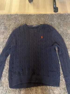 Mörkblå kabelstickad tröja Polo Ralph Lauren - Kabelstickad mörkblå tröja från Polo Ralph Lauren med rund halsringning och den klassiska röda logotypen broderad på bröstet. Tröjan har långa ärmar och ribbade muddar vid ärmslut och nederkant. Perfekt för lager på lager och stilren vardagslook. L