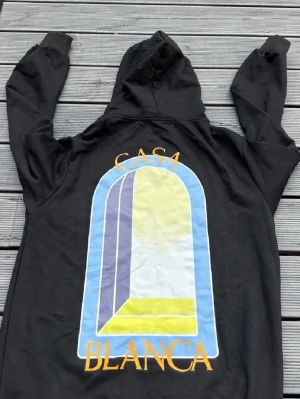 Svart hoodie från Casablanca - Säljer en svart hoodie från Casablanca med stor färgglad grafisk print och texten 'CASABLANCA' på ryggen samt mindre tryck på bröstet. Hoodien har huva och känguruficka framtill. Tillverkad i mjuk bomull, perfekt för en chill och stilren look.