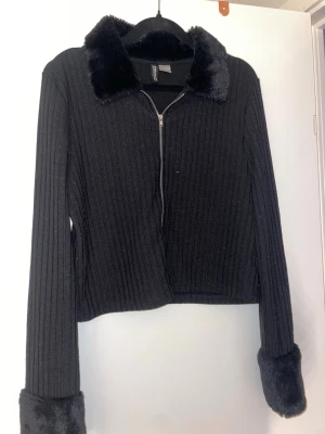 Svart ribbad jacka med fuskpälsdetaljer - Snygg svart ribbad jacka från h&m med dragkedja framtill. Jackan har mjuk fuskpäls på krage och ärmslut, vilket ger en cool och trendig look. Croppad modell med lång ärm, perfekt till höst och vinter.
