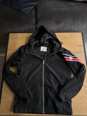 Svart vindjacka från Moncler - Svart vindjacka från Moncler med huva och hel dragkedja. Jackan har en röd och vit rand med Moncler-logga på ena ärmen. Materialet är tunt och smidigt, perfekt för blåsiga dagar. Elastiska muddar vid ärmslut och justerbar huva.