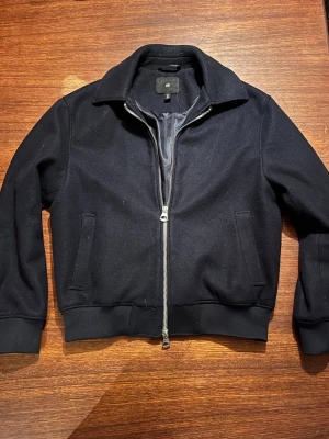 Mörkblå bomberjacka från H&M - Snygg mörkblå bomberjacka från H&M med klassisk krage och ribbade muddar. Jackan har två sidofickor och är tillverkad i ett mjukt material som känns bekvämt. Dragkedjan är lite slö, men den fungerar. Perfekt för dig som vill ha en stilren och enkel jacka till vardags.
