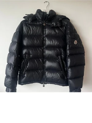 Svart dunjacka från Moncler  - Snygg svart dunjacka från Moncler med glansig finish, hög krage och dragkedja framtill. Jackan har två snedställda fickor med dragkedja och en ficka med lock och Moncler-logga på ärmen. Perfekt för kalla vinterdagar