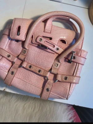 Rosa croco-mönstrad handväska - Cool rosa handväska med croco-mönster och silvriga metalldetaljer. Väskan har dubbla handtag och flera dekorativa remmar med nitar och spännen. Perfekt för dig som vill sticka ut med en trendig accessoar.