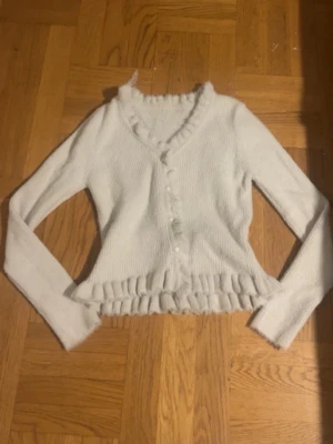 Vit ribbad kofta med volangkant - Söt vit kofta med ribbad struktur och volangkant längs nederdelen och halsringningen. Koftan har pärlknappar framtill och långa ärmar. Perfekt att styla med jeans för en trendig look. Jag skulle tro koftan passar både Xs och S.