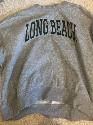 Collage tröja - Säljer en grå sweatshirt med texten LONG BEACH i stora svarta bokstäver på bröstet. Tröjan har rund halsringning, ribbade muddar och är tillverkad i mjukt bomullsmaterial. Perfekt för en avslappnad och sportig stil.
