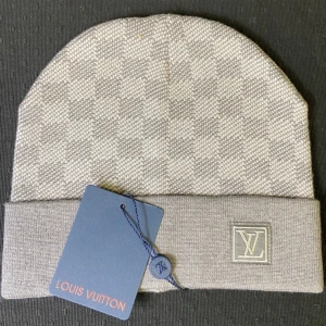Lv mössa - Snygg grå mössa från Louis Vuitton med klassiskt rutmönster och vikt kant. LV-logga i silver på framsidan ger en exklusiv touch. Tillverkad i mjukt stickat material som håller dig varm och stilren under kalla dagar.