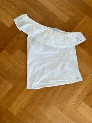 Vit oneshoulder topp med volang - Säljer en vit oneshoulder topp med bred volangkant i broderad spets. Toppen har en axel och är tillverkad i mjukt bomullstyg. Perfekt för sommarens alla tillfällen och ger en fräsch och trendig look.