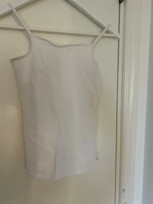 Vit enkel linnetopp med smala band - Säljer en vit linnetopp med smala axelband och rak passform. Toppen är enkel och stilren, perfekt att matcha med olika outfits. Tillverkad i mjuk bomull för en bekväm känsla.