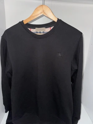Svart sweatshirt från Burberry - Säljer en svart sweatshirt från Burberry med rund halsringning och diskret broderad logga på bröstet. Tröjan har klassisk passform och är tillverkad i mjukt bomullsmaterial. Perfekt för en clean och stilren look. Storleken är M men kan passa oxkså för storlek S eftersom jag är storlek S och det passa mig också. Finns bild också!