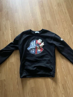 Svart Moncler sweatshirt med Popeye - Cool svart sweatshirt från Moncler med stort Popeye-tryck på bröstet och Moncler-logga på ärmen. Tröjan har rund hals och ribbade muddar. Perfekt för dig som gillar streetwear och unika prints.