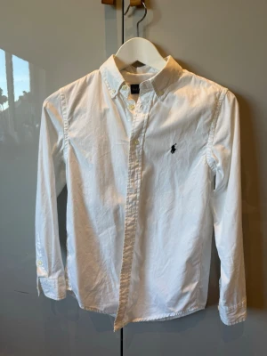 Vit skjorta från Ralph Lauren - Klassisk vit skjorta från Ralph Lauren med broderad logga på bröstet. Skjortan har lång ärm, knappar framtill och krage. Tillverkad i bomull för en skön och stilren look.