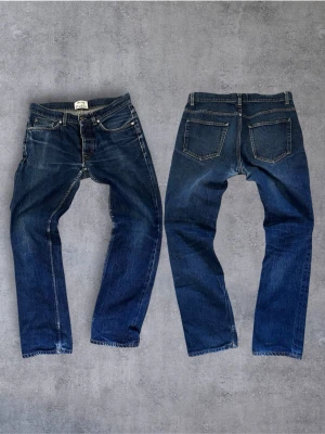  Acne Studios jeans med fet tvätt  - Säljer ett par klassiska mörkblå jeans från Acne Studios med raka ben och femficksmodell. Jeansen har snygga slitningar och kontrastsömmar, tillverkade i kraftig denim. Perfekta för en avslappnad och trendig look.