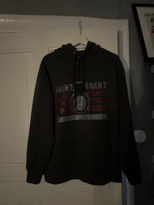  Saint Laurent hoodie - Snygg hoodie från Saint Laurent med stort tryck framtill i vitt och rött. Klassisk huva med dragsko och känguruficka. Perfekt för dig som vill ha en streetwear-look med exklusiv känsla. Materialet känns mjukt och bekvämt.