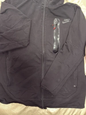 Svart Nike hoodie med dragkedja - Svart hoodie från Nike med hel dragkedja framtill och huva. Bröstet har en snygg Nike-logga och en vertikal ficka med vattentät dragkedja och röda detaljer. Tillverkad i mjukt material, perfekt för en sportig och avslappnad stil.