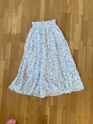 Vit långkjol med blå blommor från Gina Tricot - Säljer en superfin vit långkjol från Gina Tricot med blått blommönster. Kjolen har en smockad midja som gör den bekväm och sitter snyggt. Materialet är mjukt och lätt, perfekt för varma dagar. Passar dig som gillar romantisk och avslappnad stil.