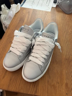 Silvriga Numeris - Snygga silvriga sneakers från NUMER16 med chunky vit sula och breda vita skosnören. Skorna har en låg profil och är tillverkade i ett glansigt material som ger en modern look. Perfekta för dig som vill sticka ut med stilrena detaljer.