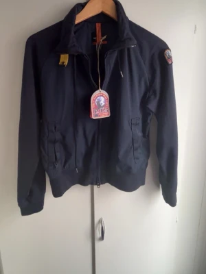 Mörkblå vindjacka från Parajumpers - Snygg mörkblå vindjacka från Parajumpers med dragkedja framtill, hög krage och två fickor med dragkedja. Jackan har en patch på ärmen och detaljer i orange. Tillverkad i ett lätt och slitstarkt material, perfekt för blåsiga dagar.