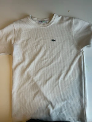 Vit ribbad t-shirt från Lacoste - Säljer en stilren vit t-shirt från Lacoste med det klassiska krokodilmärket broderat på bröstet. T-shirten har ribbad struktur, rund halsringning och korta ärmar. Perfekt för dig som gillar enkel och clean stil.