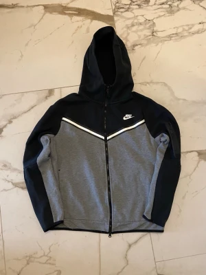 Nike tech fleece svart och grå-S - Nike tech fleece med dragkedja i svart och grått. Tröjan har huva, vita detaljer över bröstet och Nike-logga på vänster sida. Tillverkad i mjukt material, perfekt för en sportig och avslappnad stil. Långa ärmar och fickor på sidorna. Storlek S/Small!