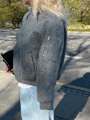 Grå bomberjacka i ullmix - Snygg grå bomberjacka med dragkedja framtill och fickor på sidorna samt en ärmficka med zip. Jackan har en mjuk, ullig textur och ribbade muddar vid ärmslut och nederkant. Perfekt för dig som gillar en avslappnad men trendig stil.