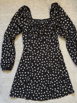 Svart blommig klänning med puffärm - Svart kortklänning med vita blommor och långa puffärmar. Klänningen har fyrkantig ringning, snörning framtill och lätt markerad midja. Perfekt för dig som gillar romantisk stil med trendiga detaljer.