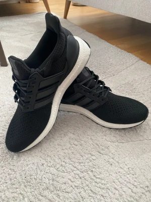 Svarta Adidas Ultra Boost sneakers - Säljer ett par svarta Adidas Ultra Boost sneakers med vit sula och klassiska tre ränder på sidan. Ovandelen är i ventilerande mesh och skon har en bekväm Boost-mellansula. Perfekt för dig som gillar stilrena och sportiga sneakers.