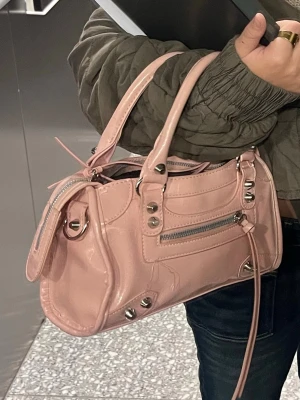 Ljusrosa handväska med silverdetaljer - Trendig ljusrosa handväska i glansigt material med silverfärgade nitar och dragkedjor. Väskan har dubbla handtag, dekorativa sömmar och en framficka med dragkedja. Perfekt för dig som gillar en cool och modern look.