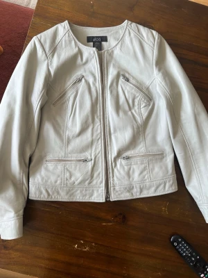 Ljusbeige skinnjacka från Ellos - Snygg ljusbeige skinnjacka från Ellos med rund halsringning och fyra dragkedjeförsedda fickor framtill. Jackan har en rak passform och stilrena sömdetaljer som ger en modern look. Perfekt för dig som vill ha en clean och trendig stil.