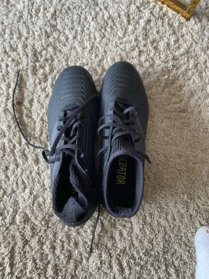 Svarta Adidas Predator fotbollsskor - Säljer ett par svarta Adidas Predator fotbollsskor med klassisk snörning och högre strumpliknande skaft. Skorna har guldtext på hälen och de ikoniska tre ränderna på sidan. Tillverkade i syntetmaterial och designade för bra grepp på planen.