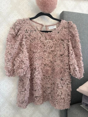 Rosa blus - Säker denna fina blus då den aldrig kommer till användning. Blusen har 3D-blommor över hela tyget, rund halsringning och puffiga trekvartsärmar. Materialet är genombruten spets med blommiga detaljer som ger en lyxig och unik look.