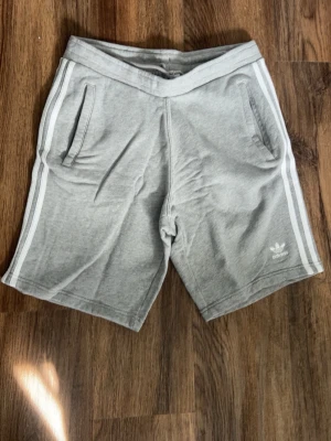 Grå Adidas shorts med vita ränder - Snygga grå shorts från Adidas med klassiska vita ränder längs sidorna och Adidas-logga på vänster ben. Shortsen har två fickor fram och är tillverkade i mjuk bomull, perfekta för en avslappnad stil.