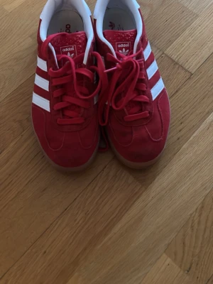 Adidas Gazelle röda sneakers - Riktigt snygga Adidas Gazelle sneakers i rött mocka med klassiska vita stripes och vit häl. Skorna har en låg profil och gummisula som ger en retro vibe. Perfekta för dig som vill sticka ut med färg och ikonisk design. Seriöst använt typ 4 gånger!