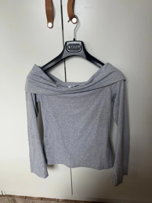 Grå offshoulder topp från Design Si - Snygg grå offshoulder topp från Design Si i mjukt material. Toppen har långa ärmar och en bred krage som vilar över axlarna för en trendig look. Perfekt att styla med jeans eller kjol för en cool outfit.