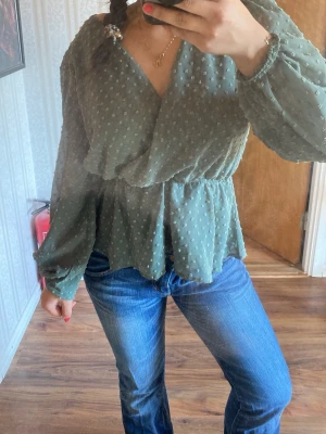 Grön omlottblus med prickig struktur - Stilren grön omlottblus med lång ärm och markerad midja. Blusen har en prickig struktur i tyget och lätt puffade axlar för en trendig look. Perfekt att matcha med jeans eller kjol för en snygg outfit.