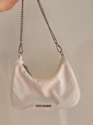 Vit axelremsväska Steve Madden - Snygg vit axelremsväska från Steve Madden i mjukt material med silverfärgade metalldetaljer och kedja. Väskan har ett minimalistiskt utseende och logga framtill. Avtagbar axelrem i tyg och matchande liten accessoarväska medföljer.