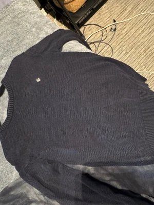 Mörkblå stickad tröja från MORRIS - Snygg mörkblå stickad tröja från MORRIS med rund halsringning och diskret broderad logga på bröstet. Tröjan har ribbade muddar vid ärmslut och nederkant, perfekt för en klassisk stil. Bra passform men ganska liten i storleken. Hör av dig vid minsta fundering 