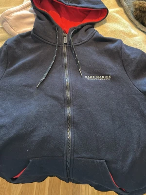 Mörkblå hoodie från Race Marine - Mörkblå zip-hoodie från Race Marine med röd insida i huvan och coola detaljer som snören och broderad logga på bröstet. Två fickor framtill och dragkedja hela vägen. Perfekt för chill dagar och snygg att matcha med jeans.