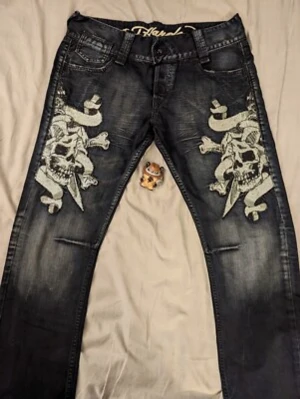Svarta jeans med dödskalletryck - Säljer ett par svarta Ed Hardy jeans med stora vita dödskalle- och benknotetryck på båda benen. Jeansen har klassisk femficksmodell, normal midja och raka ben. Materialet är denim med lätt tvättad effekt och coola detaljer.
