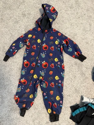 Skaloverall med huva - Mysig overall i mörkblått med färgglada tryck av Elmo, Kakmonstret och Big Bird från Sesame Street. Dragkedja framtill, muddar vid ärmar och ben samt huva. Perfekt för chill dagar eller lek utomhus.