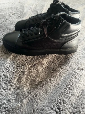 Svarta high top sneakers med dragkedja - Snygga svarta high top sneakers med dragkedja på sidan och snörning framtill. Skorna har en mix av läder och textilmaterial som ger en cool look. Perfekta för dig som gillar streetstyle och vill ha något som sticker ut lite extra.