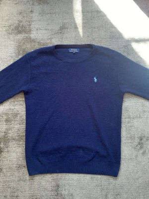 Mörkblå stickad tröja Polo Ralph Lauren - Mörkblå stickad tröja från Polo Ralph Lauren med rund halsringning och diskret strukturmönster. Tröjan har det klassiska broderade logot på bröstet i ljusblått och ribbade muddar vid ärmslut och nederkant. Perfekt för lager på lager under kyliga dagar.