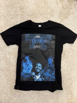 Svart Psychodrama Dave t-shirt - Säljer en svart t-shirt med tryck från Dave's album Psychodrama på framsidan. T-shirten har korta ärmar och en klassisk passform. Trycket är i blåa toner och ger en riktigt snygg streetwear-vibe.