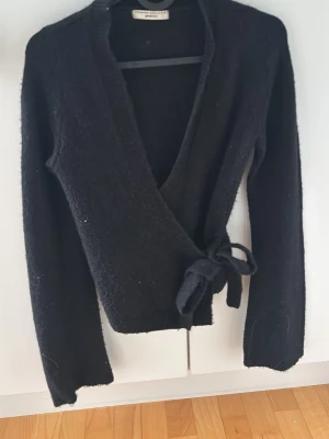 Svart omlottkofta från Gina Tricot - Svart stickad omlottkofta från Gina Tricot med knytband i midjan och djup v-ringning. Koftan har långa ärmar och ribbade kanter. Perfekt för lager-på-lager och enkel att styla till olika looks.